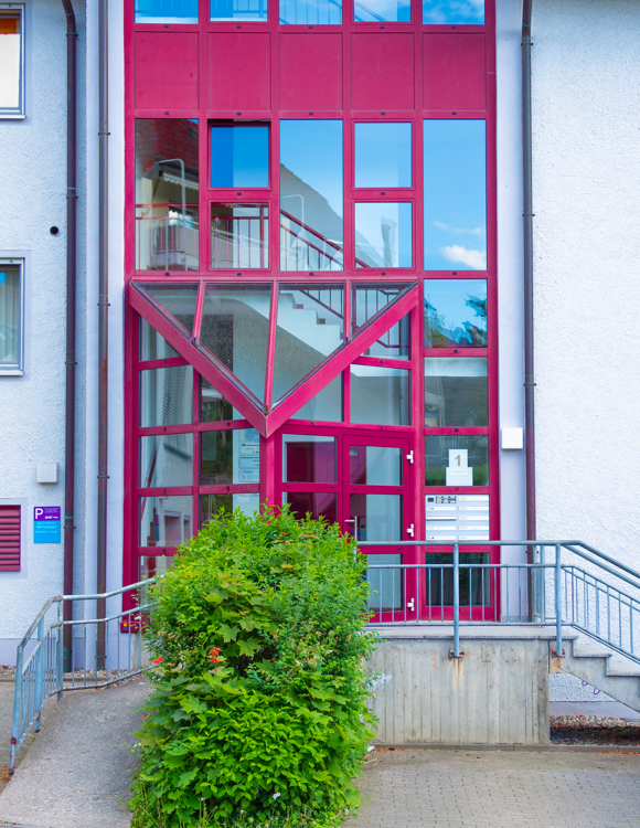 Hauseingang der Praxis mit zahlreichen rotumrahmten Fenstern und Türen